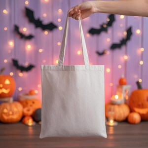 May include: A blank, off-white tote bag with long handles is held up against a Halloween-themed background. The background features carved pumpkins, string lights, and bat silhouettes, creating a festive atmosphere.