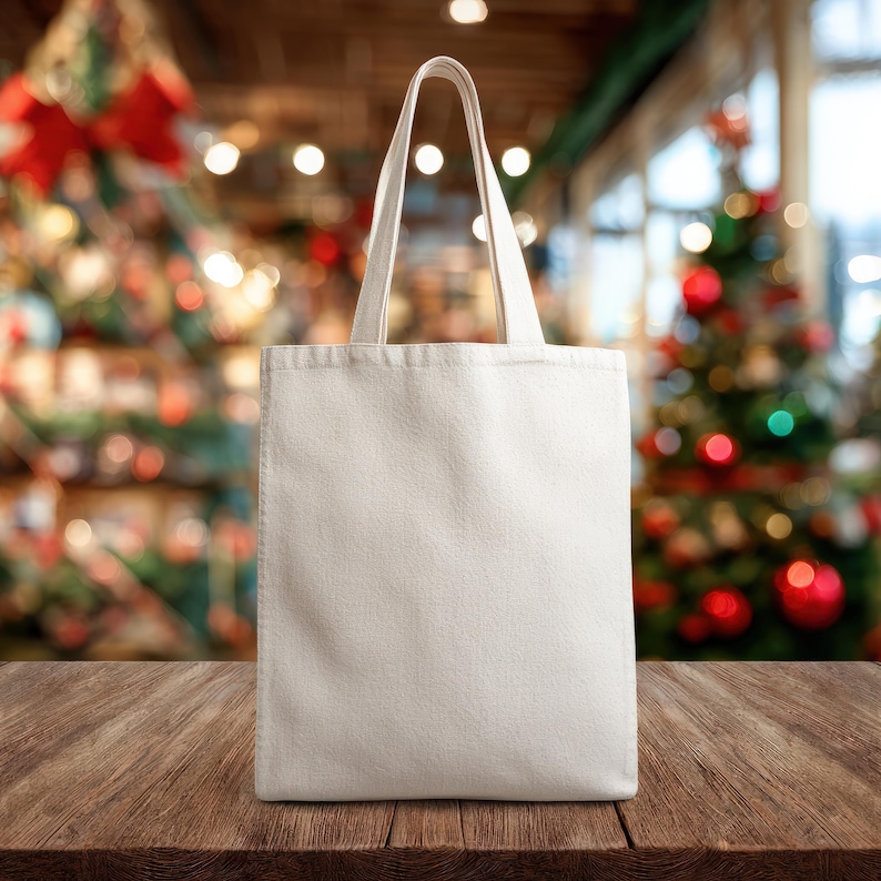 May include: A blank, off-white canvas tote bag with two handles stands on a wooden surface. The background is blurred, featuring Christmas decorations with red and green accents, creating a festive atmosphere.