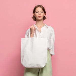 May include: A white tote bag with a rectangular shape and two handles is held by a person. The bag is made of a plain, unadorned fabric. The background is a solid pink color.