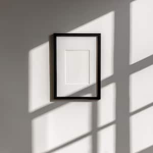 May include: A black picture frame with a white mat is mounted on a white wall. The frame is rectangular and has a simple, modern design. Sunlight casts shadows across the wall, adding depth to the image. The frame is empty, ready for a photo or artwork.