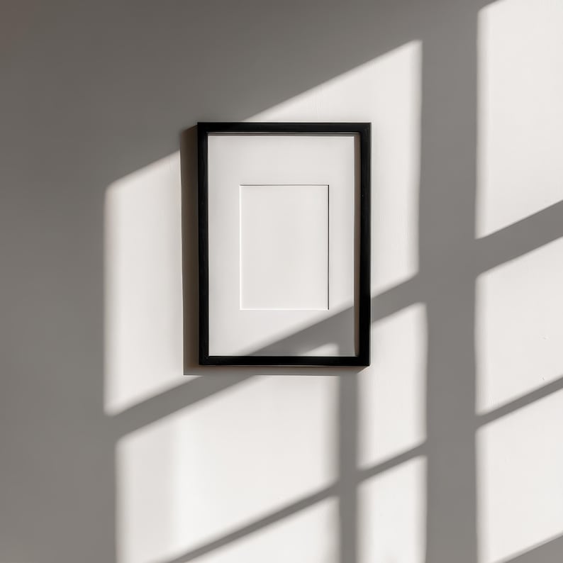 May include: A black picture frame with a white mat is mounted on a white wall. The frame is rectangular and has a simple, modern design. Sunlight casts shadows across the wall, adding depth to the image. The frame is empty, ready for a photo or artwork.
