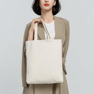 May include: A cream-colored tote bag with a rectangular shape and two handles. The bag is held by a person wearing a tan blazer and a white shirt. The background is a solid light gray.