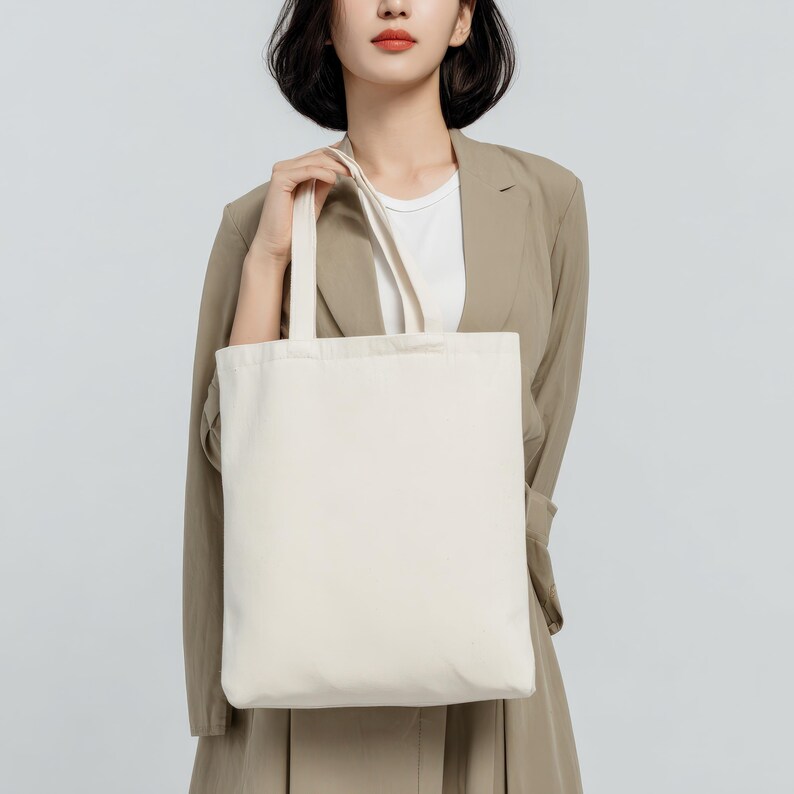 May include: A cream-colored tote bag with a rectangular shape and two handles. The bag is held by a person wearing a tan blazer and a white shirt. The background is a solid light gray.
