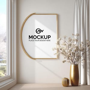 May include: A gold-framed picture frame with a white interior, displaying the word "MOCKUP" in black text. A gold vase with white flowers and a small gold decorative object are on a wooden surface.