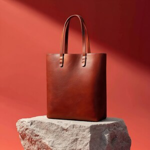 May include: A brown leather tote bag with two handles. The bag is rectangular and sits on a stone surface against a red background. The handles are attached to the bag with metal hardware.