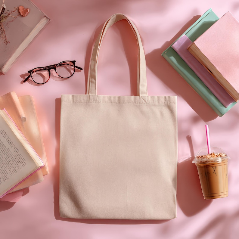 May include: A beige canvas tote bag with handles, surrounded by books, eyeglasses, and a cold coffee drink with a pink straw, all on a pink surface. The tote bag is empty and ready for use.