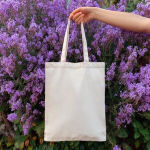 May include: A blank, cream-colored tote bag with long handles is held up against a backdrop of vibrant purple flowers. The bag is made of a sturdy fabric, perfect for carrying groceries or other items.