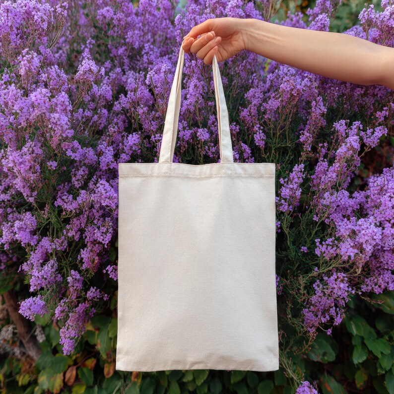 May include: A blank, cream-colored tote bag with long handles is held up against a backdrop of vibrant purple flowers. The bag is made of a sturdy fabric, perfect for carrying groceries or other items.