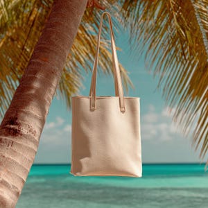 May include: A beige tote bag with long, thin straps hangs from a palm tree. The bag has a rectangular shape and a simple design. The background features a tropical beach scene with turquoise water and a blue sky.