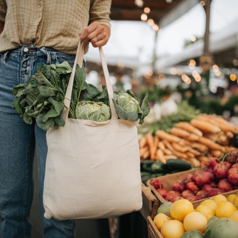May include: A beige canvas tote bag filled with fresh green vegetables, including leafy greens and cabbages, is held by a person at a farmer's market. The background shows a variety of produce, including carrots, beets, and lemons.