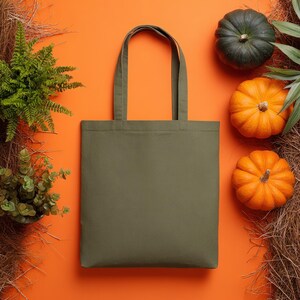 May include: Olive green tote bag with matching handles, set against an orange background. The bag is surrounded by green plants, two orange pumpkins, and a dark green pumpkin, suggesting a fall theme.