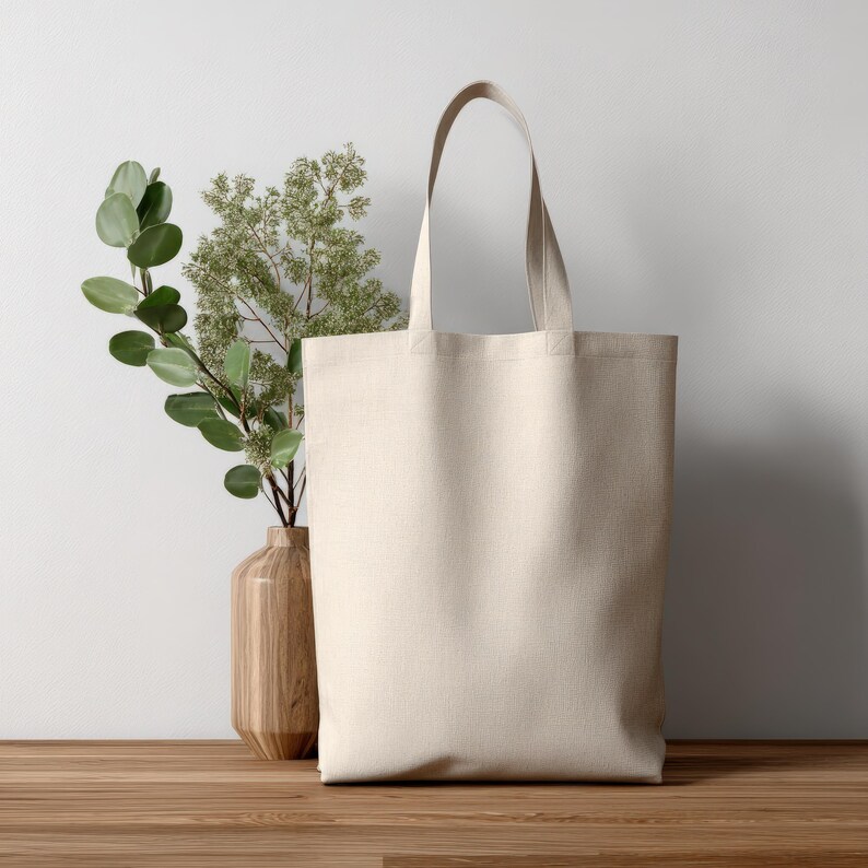 May include: A blank, cream-colored tote bag with long handles stands on a wooden surface. A wooden vase with green and light green leafy branches is to the left of the bag. The background is a white wall.
