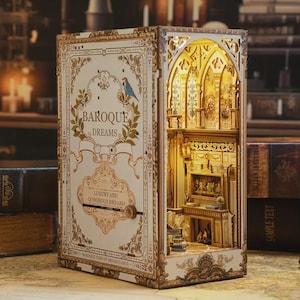 May include: A decorative wooden book-shaped diorama with the text "Baroque Dreams." The interior features a detailed miniature baroque-style room with a fireplace and arched windows, illuminated by warm lighting. The exterior is cream-colored with gold accents.