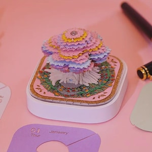 May include: A detailed paper art sculpture of a tree in pastel shades of pink, yellow, and purple. The artwork is set on a decorative base with a gold glitter trim, inside a white square box. The word "Janwary" is visible.