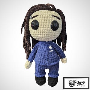 May include: A handmade crochet doll with dark brown yarn hair, large black plastic eyes, and a light beige face. The doll wears a blue textured yarn tracksuit with a white embroidered logo on the chest and black shoes. Includes a Memento Mori by Julz logo.