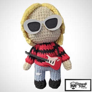 May include: Crocheted doll of a musician with blonde hair, white sunglasses, and a red and black striped sweater. The doll is holding a red guitar and wearing blue jeans. The doll is a handmade item.