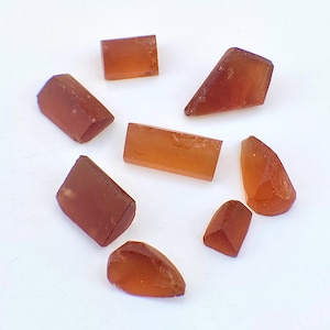 May include: A selection of raw, orange-brown gemstones arranged in a circular pattern. The stones vary in shape, with some appearing rectangular and others more triangular. The gemstones are translucent, allowing light to pass through.
