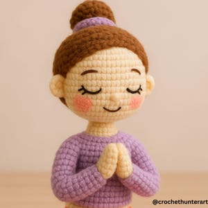 May include: A crocheted doll with brown hair styled in a bun, closed eyes, and rosy cheeks. The doll wears a lavender sweater and has hands clasped together in a prayer position. The doll is a handmade item.