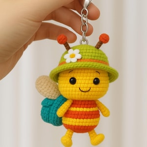 May include: A handmade crochet bee keychain. The bee is yellow with orange stripes, black eyes, and a smiling mouth. It wears a green hat with a white flower and has small orange antennae. The bee has teal wings and yellow feet.