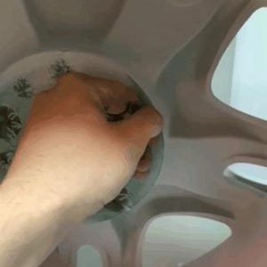 Op de afbeelding: Een hand houdt een helder, cirkelvormig object met een bloemenpatroon in een lichtgrijze, wielvormige structuur. De structuur heeft meerdere openingen en een glad oppervlak.