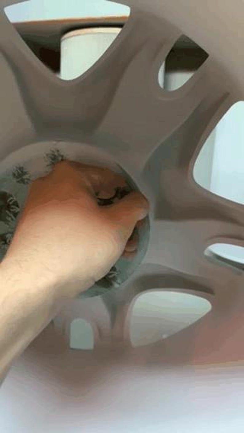 Op de afbeelding: Een hand houdt een helder, cirkelvormig object met een bloemenpatroon in een lichtgrijze, wielvormige structuur. De structuur heeft meerdere openingen en een glad oppervlak.