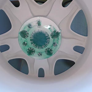 Op de afbeelding: Een wit, cirkelvormig object met een reeks gebogen uitsparingen en een centraal, cirkelvormig ontwerp. Het centrale ontwerp heeft een groen, getextureerd patroon met kleine, stekelige details. Het object lijkt een onderdeel van een groter apparaat te zijn.