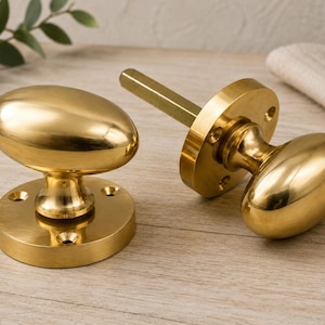May include: Two polished brass door knobs with an oval shape and a smooth, reflective finish. Each knob is attached to a circular mounting plate featuring four pre-drilled screw holes. One knob includes a square metal spindle extending from the back.