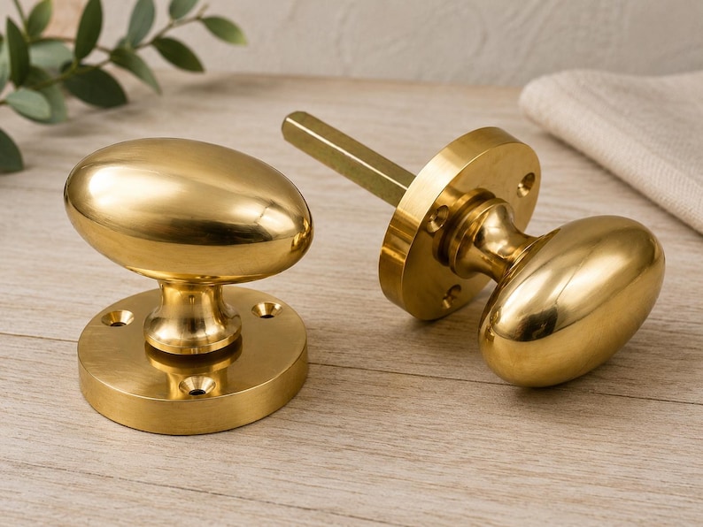 May include: Two polished brass door knobs with an oval shape and a smooth, reflective finish. Each knob is attached to a circular mounting plate featuring four pre-drilled screw holes. One knob includes a square metal spindle extending from the back.