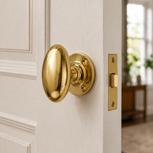 May include: A polished brass oval door knob with a matching circular rosette plate and a rectangular latch plate, installed on a white paneled door. The hardware features a smooth, reflective metallic finish and visible mounting screws.