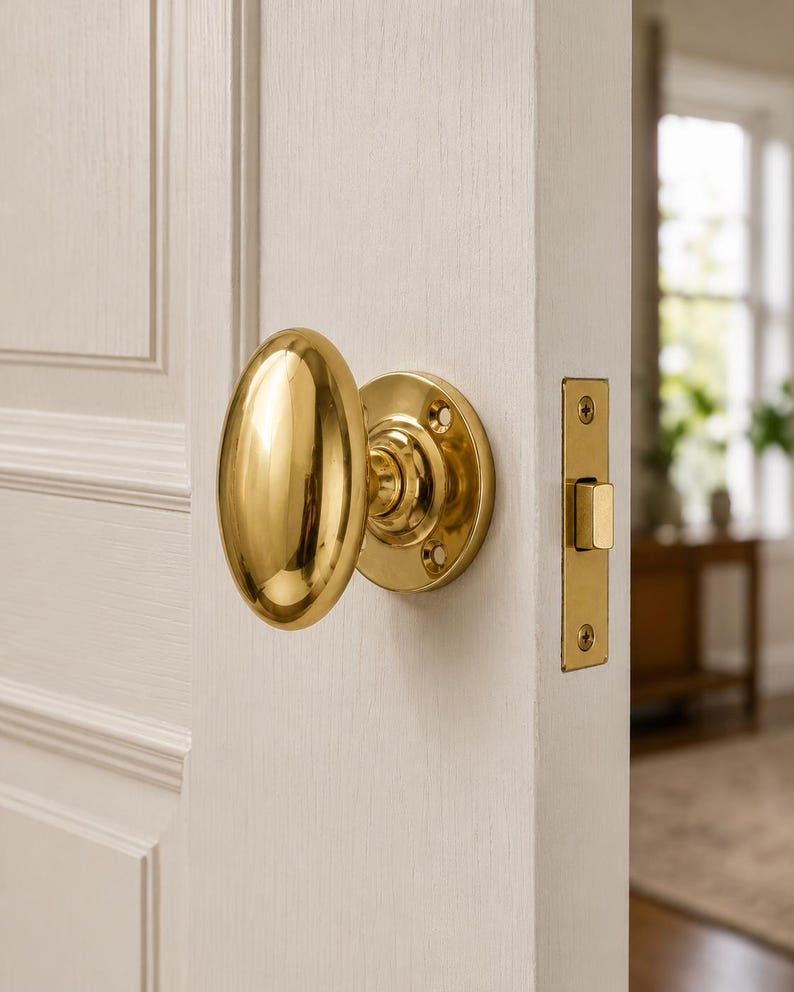 May include: A polished brass oval door knob with a matching circular rosette plate and a rectangular latch plate, installed on a white paneled door. The hardware features a smooth, reflective metallic finish and visible mounting screws.