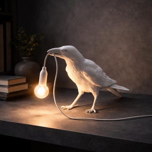 May include: A white crow-shaped lamp, holding a lightbulb in its beak. The lamp is on a dark surface, with the lightbulb lit. The lamp is a decorative item, and the cord is visible.