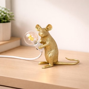 May include: A gold-coloured mouse figurine holding a light bulb. The mouse is in a standing position, with its front paws holding the white light socket. The light bulb is clear with a yellow filament. A white cord extends from the light socket.