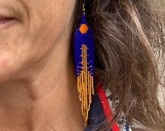 May include: A pair of long, beaded earrings with a blue and gold color scheme. The earrings are made of small, round beads that are strung together in a geometric pattern. The top of the earrings features a small, orange bead.