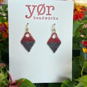 May include: A pair of handmade beaded earrings. Each earring is a diamond shape, with a top section in red and a bottom section in black and gray. The earrings hang from gold-colored hooks. The card behind the earrings reads "yør beadworks".