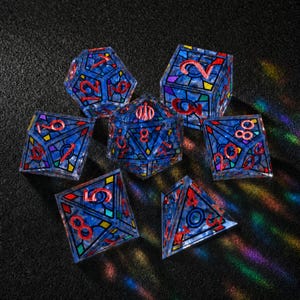 May include: A set of seven polyhedral dice featuring a translucent blue base with a stained-glass pattern of black lines and colorful geometric accents. Each die has engraved numbers filled with a bright red pigment for high visibility.