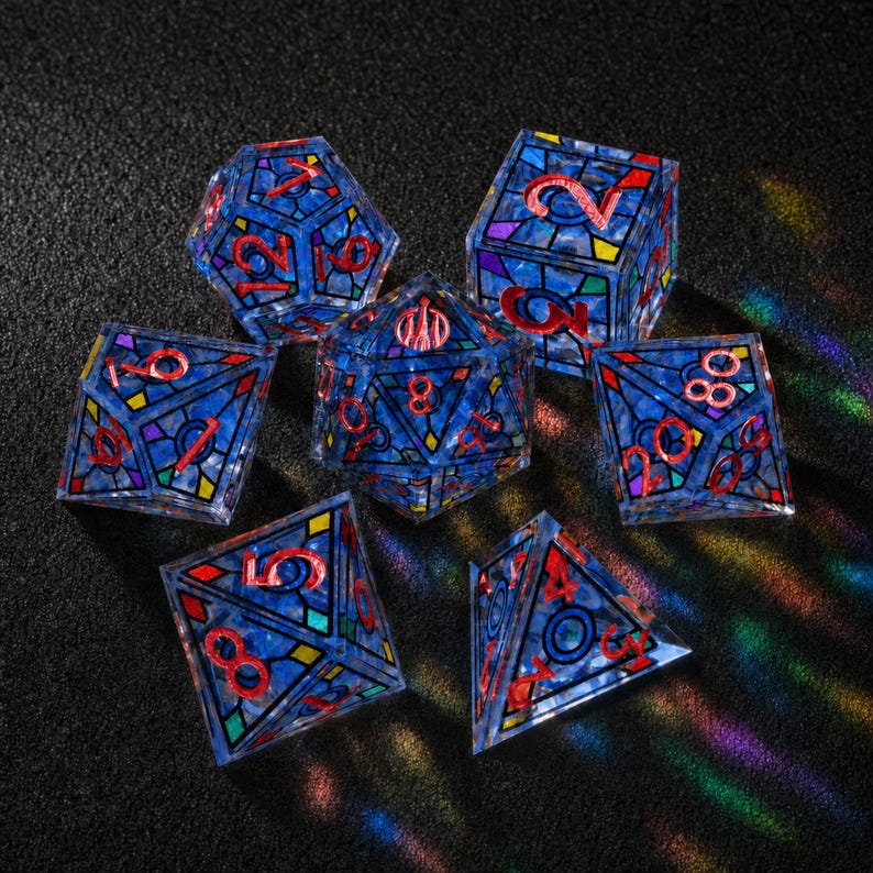 May include: A set of seven polyhedral dice featuring a translucent blue base with a stained-glass pattern of black lines and colorful geometric accents. Each die has engraved numbers filled with a bright red pigment for high visibility.
