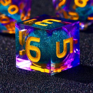 Può includere: Primo piano di un dado a sei facce con numeri dorati incastonati in resina trasparente sfumata di viola. L'interno ha un nucleo glitterato color verde acqua. I numeri sono grandi e facili da leggere, con un focus sul numero sei.