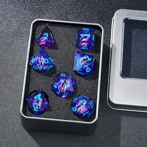 Könnte beinhalten: Ein Satz von sieben polyedrischen Würfeln in einer Metalldose. Die Würfel sind transparent mit einem wirbelnden lila und blauen Design und blauen Zahlen. Die Würfel haben verschiedene Formen, darunter ein d4, d6, d8, d10, d12, d20 und ein Prozentwürfel.
