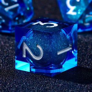 Puede incluir: Primer plano de un dado azul transparente de seis caras con el número dos en plata. El dado tiene un interior azul brillante. El número tres es visible en la parte superior del dado. El fondo es oscuro.