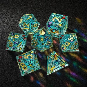 May include: A set of seven polyhedral dice featuring a stained-glass aesthetic with translucent teal, blue, and purple segments outlined in black. Each die has gold-colored numbers etched into the surface, including a d20, d12, d10, d8, d6, and d4.