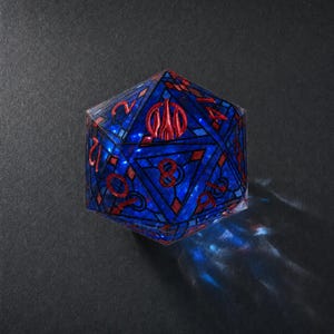 May include: A twenty-sided die featuring a deep blue translucent body with a stained glass-like pattern of dark blue lines. The numbers and a central emblem are embossed in a metallic red finish, creating a sharp contrast against the blue.