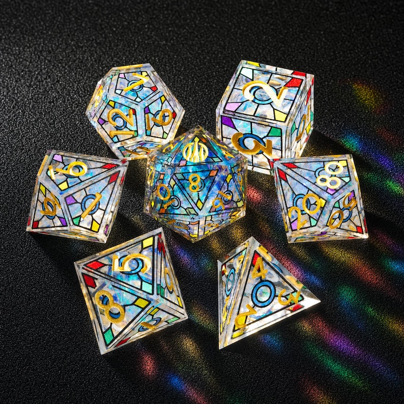 May include: A set of seven polyhedral dice featuring a stained glass pattern with translucent blue, purple, and green segments outlined in black. Each die has gold-colored numbers etched into the surface, reflecting light with a prismatic effect.