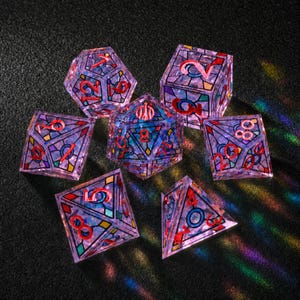 May include: A set of seven polyhedral dice featuring a translucent purple base with a stained-glass pattern of black lines and colorful geometric shapes. Each die has bright red numbers etched into the surface, creating a vibrant contrast.
