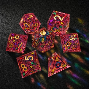 May include: A set of seven polyhedral dice featuring a stained-glass aesthetic with translucent pink, purple, and green geometric segments outlined in black. Each die face displays gold-colored numbers in a clear, legible font style.
