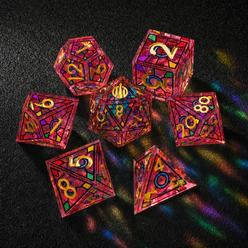 May include: A set of seven polyhedral dice featuring a stained-glass aesthetic with translucent pink, purple, and green geometric segments outlined in black. Each die face displays gold-colored numbers in a clear, legible font style.