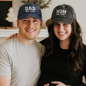 Może przedstawiać: Para nosi pasujące czapki z daszkiem. Granatowa czapka ma napis "DAD EST. 2025" białymi literami. Druga czapka jest szara i ma napis "MOM EST. 2025" białymi literami. Kobieta jest w ciąży.