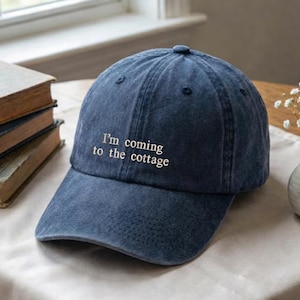 May include: A navy blue baseball cap with the embroidered text "I'm coming to the cottage". The cap is made of a soft, textured fabric and has a curved brim. The cap is sitting on a table next to a stack of books and a small vase of flowers.