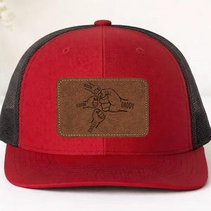 May include: Red and black trucker hat with a brown leather patch. The patch features a graphic of four fists bumping, with the words "Daddy", "Colton", "Kinsley", and "Jackson" printed on it. The hat has a curved brim and a mesh back.