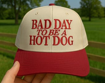 May include: A baseball cap with a cream-colored crown and a red brim. The cap features the embroidered phrase "BAD DAY TO BE A HOT DOG" in red lettering. The cap is a classic six-panel design.