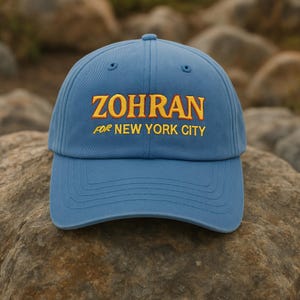 Könnte beinhalten: Blaue Baseballkappe mit goldfarben gesticktem Schriftzug "ZOHRAN" und darunter "FOR NEW YORK CITY". Die Kappe hat einen gebogenen Schirm und ist aus weichem Stoff gefertigt. Ein lässiges Accessoire.